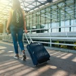 An image of a woman wearing a backpack and pulling a small suitcase on wheels.